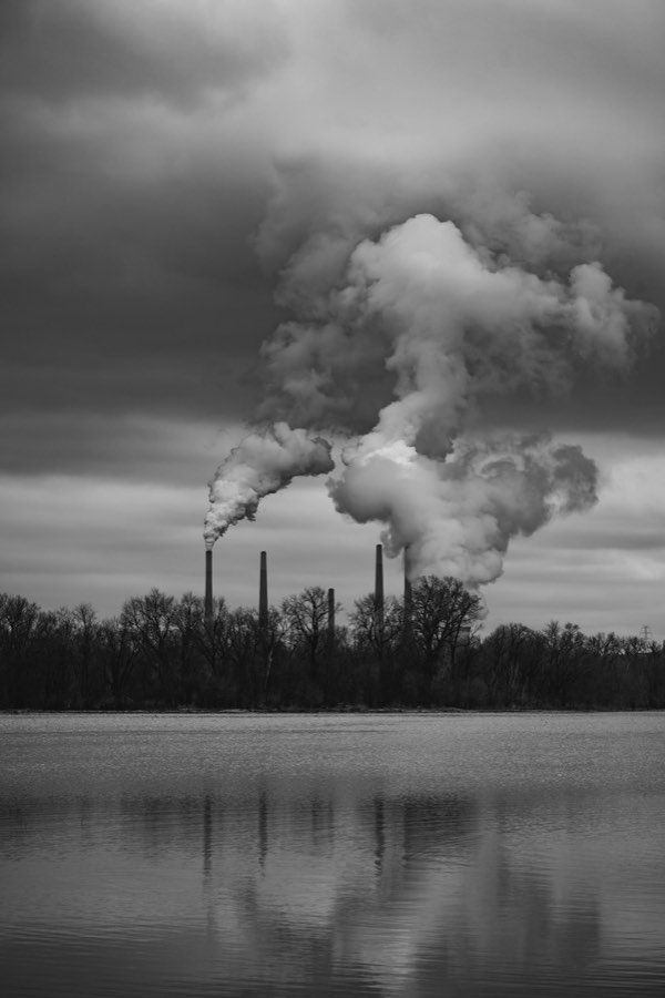Black-and-white photograph of power-station smokestacks releasing plumes over a treeline and lake